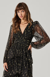 A woman wearing a black, semi-sheer dress with a sparkly, metallic floral pattern. The dress has a V-neckline, elastic waist, and long, billowy sleeves gathered at the wrists. Her hair is long and wavy with bangs, and she has a neutral expression.